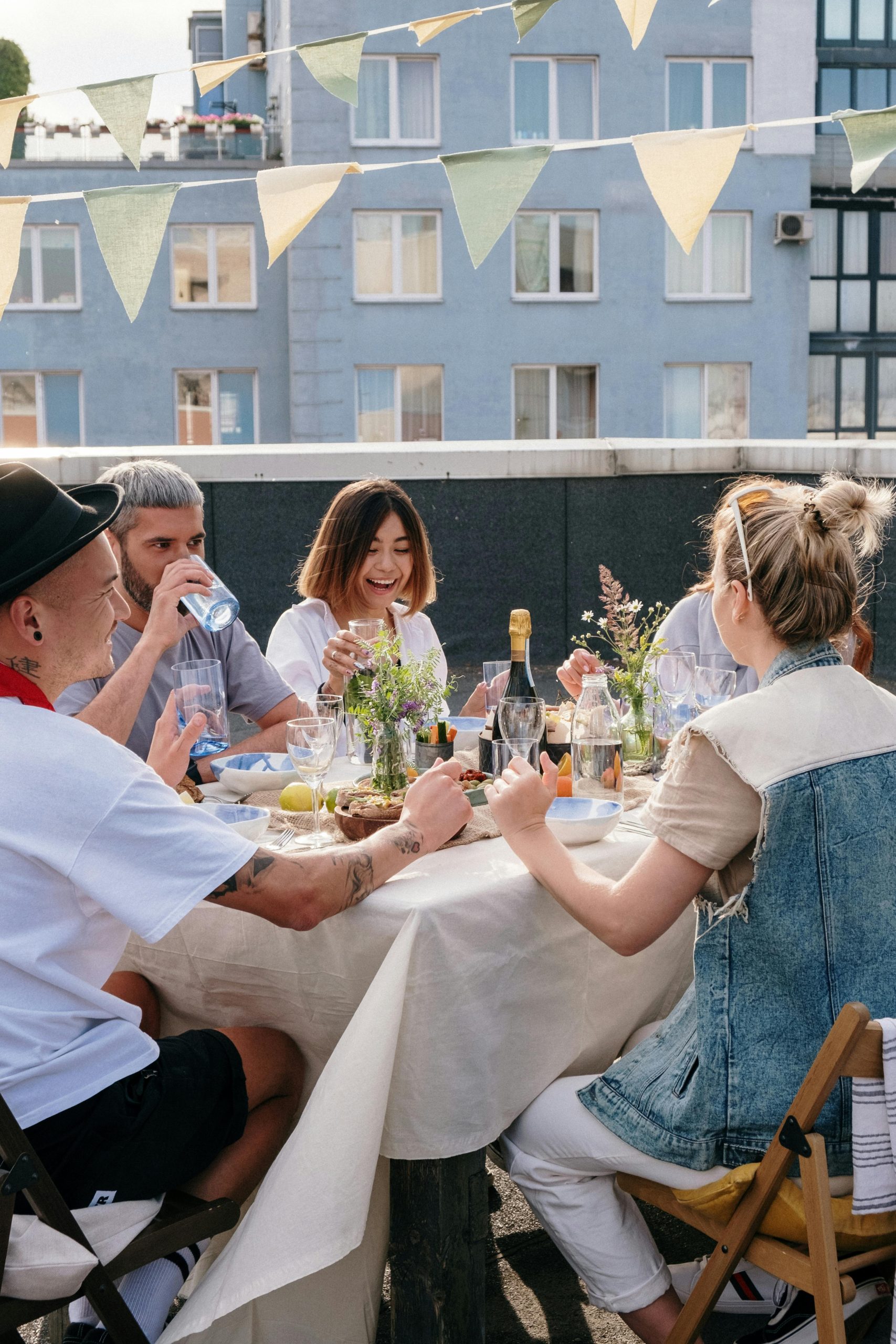 Friends enjoy a casual rooftop meal with drinks and laughter under bunting flags.